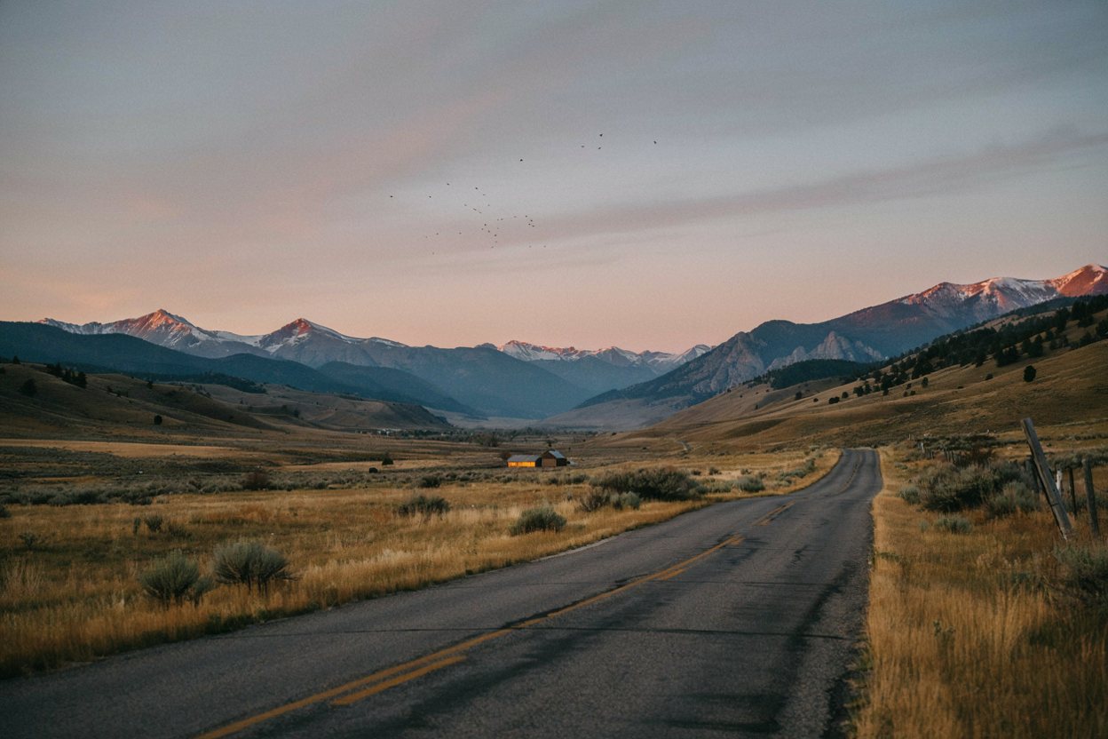 A two-lane road at dawn leading through Colorado foothills toward snow-capped mountains, a farmhouse light glowing in the valley